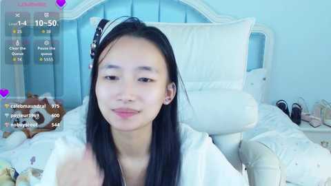Video of an Asian woman with long black hair, wearing a white robe, sitting on a bed with white pillows and a headboard.
