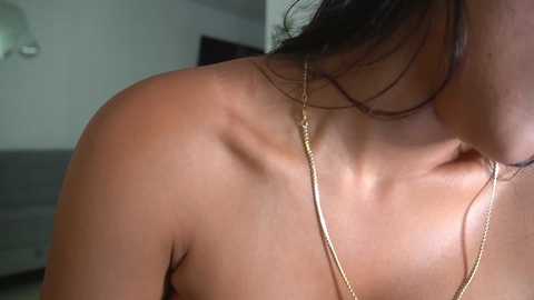 A close-up video of a topless woman with medium brown skin, wearing a gold necklace, and long dark hair. The background is blurred, showing a grey couch and white wall.