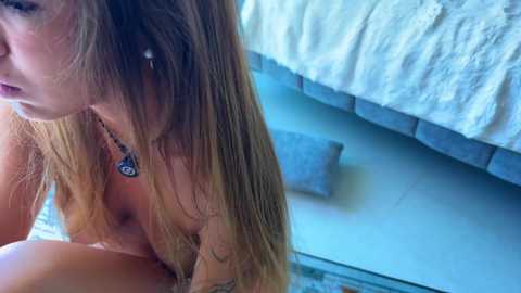 Media: A video of a nude woman with long brown hair, sitting on a tiled floor, wearing a necklace. The background features a bed with a white quilt and gray pillows.