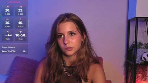 Media: Video of a young woman with long brown hair, wearing a black top, sitting indoors. Background shows a temperature display, a plant, and a lamp.