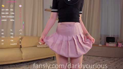 Media: Video of a slender, fair-skinned person with a black shirt and pleated pink skirt, standing in a modern living room with beige curtains and a yellow sofa.