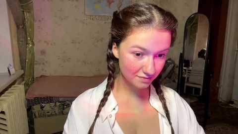 A video of a young Caucasian woman with long brown hair in braids, wearing a white shirt, standing in a dimly lit, vintage bedroom with floral wallpaper and a bed with a patterned quilt.