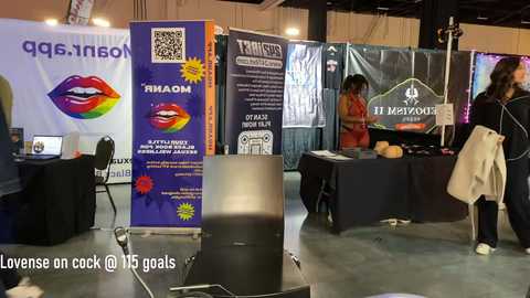 Video of a trade show booth featuring a large \"Loves on Cock\" sign, LGBT-themed posters, and a woman in a red dress at a table, with a beige coat draped over a chair.