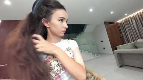 Video of a young woman with long, voluminous brown hair styled in a high ponytail. She is wearing a white t-shirt with a cartoon character. The background features a modern, minimalist living room with white walls, wooden furniture, and a staircase.