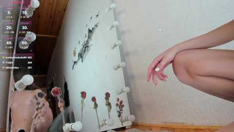 A video shows a close-up of a woman's bare legs, partially visible in a dimly lit room with a large, reflective mirror adorned with white bulbs.