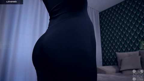 Video of a woman in a tight, black dress, accentuating her curves, standing in a modern bedroom with diamond-patterned wallpaper and a beige couch.