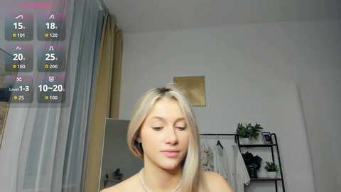 Media: Video of a blonde woman with light skin, wearing a white top, standing in a modern, minimalistic bedroom with a mirror, shelves, and curtains.