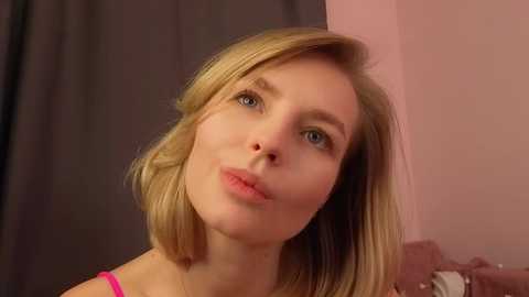 A video of a young Caucasian woman with straight, shoulder-length blonde hair, light skin, and blue eyes, wearing a pink top, indoors against a muted pink and dark grey background.