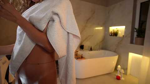 A video of a partially nude woman with a towel covering her chest, standing in a modern bathroom with a white marble sink, a bathtub, and candles.