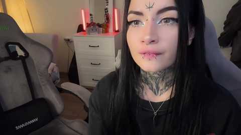 Video of a young woman with black hair, multiple piercings, and intricate tattoos, seated on a gaming chair in a dimly lit room with a white dresser and red candles.