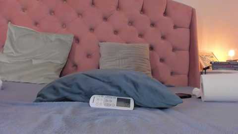 Video of a cozy bedroom with a pink tufted headboard, light blue bedspread, a blue pillow, a gray pillow, and a white alarm clock on a nightstand.