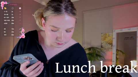 Media: A video of a young woman with blonde hair in a black top, holding a smartphone, looking at a calendar app. Background features a potted plant, wall decor, and \"Lunch break\" text.