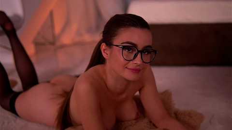 A video of a fair-skinned woman with brown hair, large black-framed glasses, and black stockings, lying topless on a bed, smiling. Background features a beige wall and a bed with white sheets.