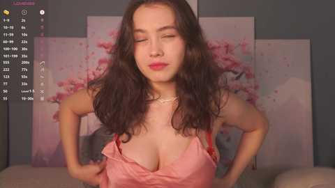 Video of a smiling Asian woman with long, wavy black hair, wearing a pink satin camisole, standing in front of a wall with floral artwork.