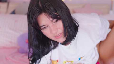 Video of a young, fair-skinned Asian woman with long black hair, wearing a white shirt, leaning forward in a softly lit bedroom with pastel decor.