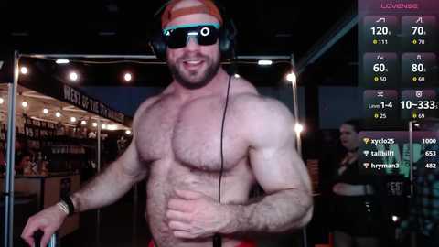 Media: Video of a shirtless, muscular man with a beard and headphones, smiling. Background includes a dimly lit room with a TV screen displaying streaming metrics.