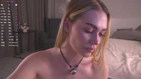 A video of a topless, fair-skinned, blonde woman with straight hair, wearing a black necklace, sitting on a bed in a dimly lit bedroom.