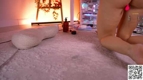 A video showing a nude woman in a dimly lit room, kneeling on a plush white carpet. A red candle and a bottle are on the floor, with a red heart-shaped cushion beside her.