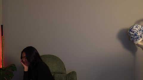Media: Video of a woman with long black hair, wearing a black sweater, sitting in a green armchair, leaning on her left hand, in a dimly lit room with beige walls, a red lamp, and a blue and white ceramic vase.