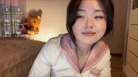A video of an East Asian woman with light skin, black hair, and a small nose. She wears a pink sailor collar top and is smiling. A teddy bear and a bed are visible in the background.