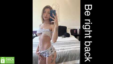 Video of a slender young woman with light skin, long blonde hair, wearing a crop top and matching shorts, taking a selfie in a bedroom. Text overlay reads: \"Be right back.\