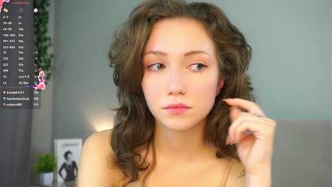 Video of a young woman with wavy brown hair, light skin, and pink lipstick, touching her chin, in a well-lit room with green walls and a potted plant.
