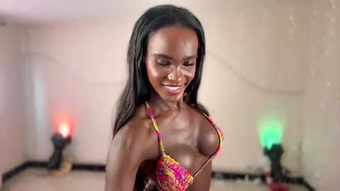 Video of a smiling, dark-skinned woman with long black hair in a colorful, revealing bikini, standing in a dimly-lit room with colored lights and a white wall.