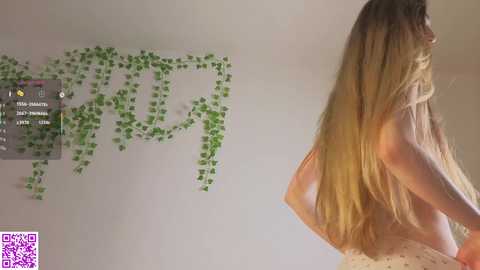 Video of a topless woman with long blonde hair, standing against a wall adorned with green ivy, in a dimly lit room.