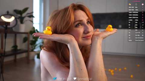 A video of a topless, fair-skinned woman with red hair, wearing yellow rubber duckies on her hands, smiling in a modern kitchen with a green plant and wooden table.