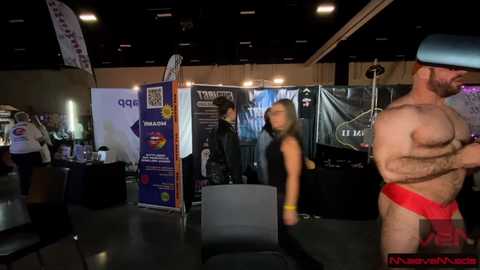 Video of a dimly-lit convention hall with a shirtless man in a red thong, a woman in a black tank top, and a man in a white shirt. Booths with banners and promotional materials in the background.