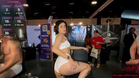 Video of a young woman with dark hair, wearing a white bikini, sitting on a chair in a dimly lit room with various people in the background.