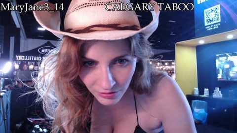 Media: Video of a Caucasian woman with wavy, light brown hair, wearing a beige cowboy hat and a black tank top, posing in a dimly lit bar, with \"MaryJane3 14\" and \"CALGARYTABS\" text overlay.