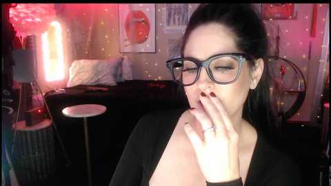 Video of a woman with long black hair, wearing black-framed glasses and a black top, sensually biting her fingers in a dimly lit, artsy room with posters and a neon light.