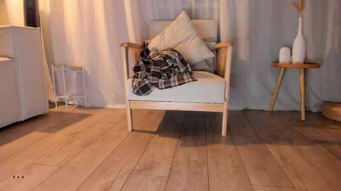 Video of a modern, light-wood armchair with white cushions, draped with a plaid blanket, set against a pale blue curtain backdrop.