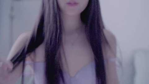 Video of a young woman with long, straight, dark hair, wearing a strapless, light purple top, standing against a soft-focus, light-colored background.