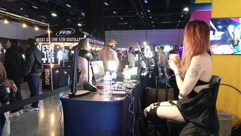 Video of a lively indoor event with a tattooed woman in a black outfit seated at a booth, holding a drink, surrounded by attendees and vendor stalls under a dimly lit, industrial-style ceiling.
