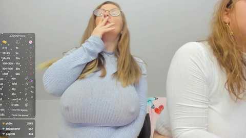 Video of a blonde woman with large breasts in a tight, light blue sweater, covering her mouth, next to a woman in a white top, in a light-colored room.