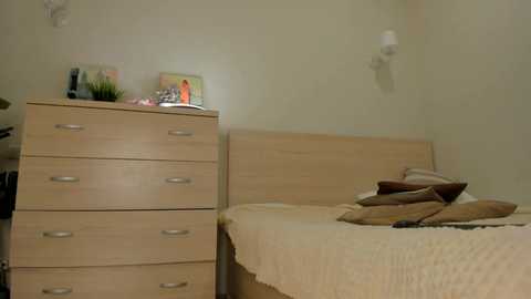 A video of a simple, sparsely decorated bedroom with a light wooden dresser, a bed with a beige quilt, and two framed pictures on top. The walls are painted a pale yellow.