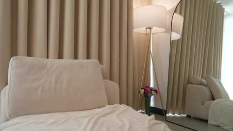 Video of a cozy bedroom with beige curtains, a plush white sofa, a bedside table with a vase of red and white flowers, and a tall, modern lamp with a white shade.