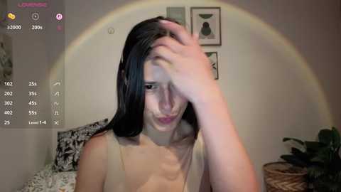 A video of a young woman with long black hair, wearing a beige top, sitting in a dimly lit room, holding her head, with a rainbow halo effect, and a smartphone screen overlay showing camera settings.