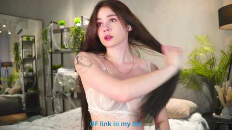 Video of a young Asian woman with long black hair, wearing a white lace bralette, standing in a modern bedroom with plants, shelves, and a gray bed.