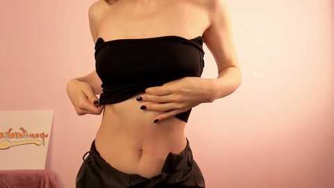 Video of a slender, light-skinned person with a small bust, wearing a black strapless top and unbuttoned jeans, standing in a pink room with a cartoon poster on the wall.