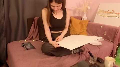 Media: A video shows a young woman with fair skin and long brown hair sitting on a pink couch, wearing a black sports bra and black leggings, drawing on white paper.