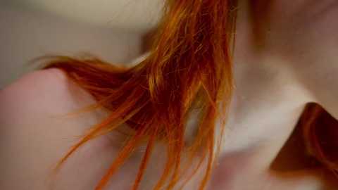 Video of a woman's shoulder and neck with vibrant red hair, partially covering her pale skin, and a white shirt collar visible. Background is blurred, focusing on the hair's texture and color.