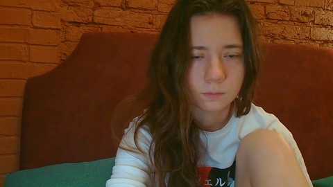Video of a young woman with long, wavy brown hair, wearing a white T-shirt, seated in a room with a brick wall and brown upholstered headboard.