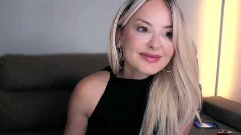 A video of a smiling, fair-skinned woman with long, straight blonde hair, wearing a black sleeveless top, sitting on a gray couch beside a lit lamp.