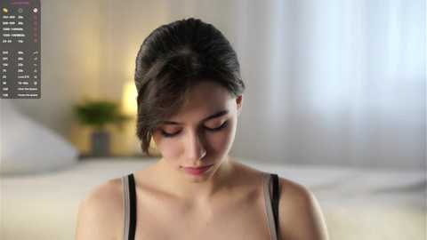 A video of a young woman with fair skin, dark hair tied back, wearing a black tank top, standing in a softly lit bedroom with blurred white curtains and a bed in the background.