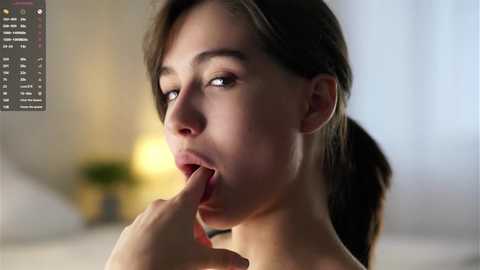Video of a young woman with light skin, brown hair, and fair complexion, seductively licking her fingers while gazing intensely at the camera, in a softly lit bedroom setting.