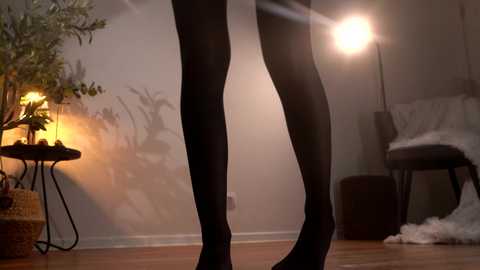 Video of a person in black leggings, standing in a softly lit room with a potted plant, a cozy chair, and a warm lamp casting shadows.