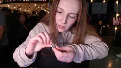 Video of a fair-skinned, long-haired woman with closed eyes, holding a small, shiny object, wearing a gray hoodie, in a dimly lit, festive indoor space with string lights and blurred background.